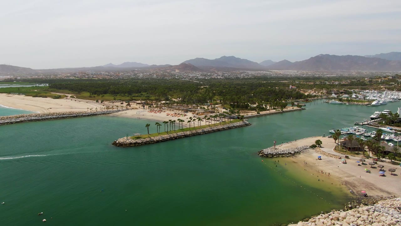 Aerial coastal inlet with boat wake, beach, palm-lined breakwater, marina docks, and mountain backdrop.