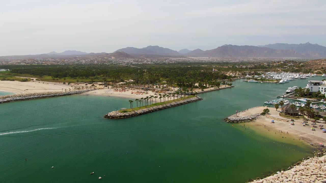 Wide aerial frame over Puerto Los Cabos Beach with green inlet water in front, sandy beach and umbrellas at the shore, marina boats near a white building, and mountains on the horizon.