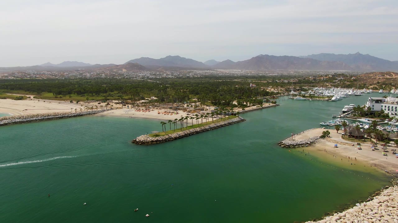 Aerial coastal inlet with a white watercraft trail crossing green water near a beach and marina.
