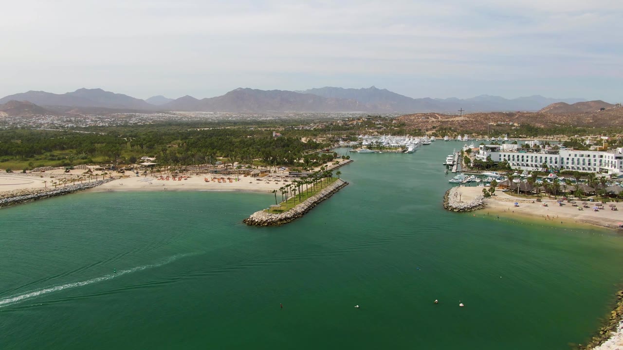 Aerial frame at Puerto Los Cabos Beach where the white wake stretches longer across the foreground water, beach umbrellas are visible along the sand, buildings and marina sit to the right, and no visible people appear.