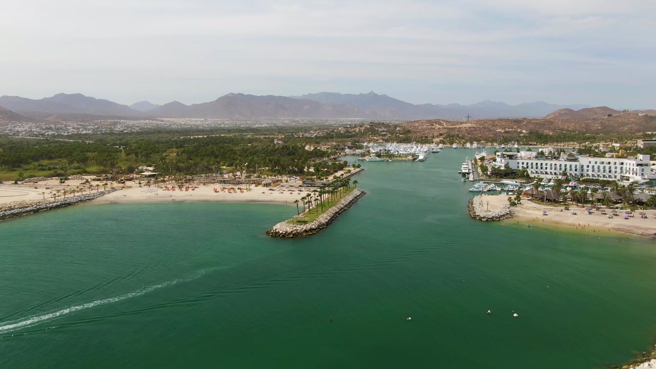Wide aerial view over calm green water toward a sandy beach, marina, and mountains.