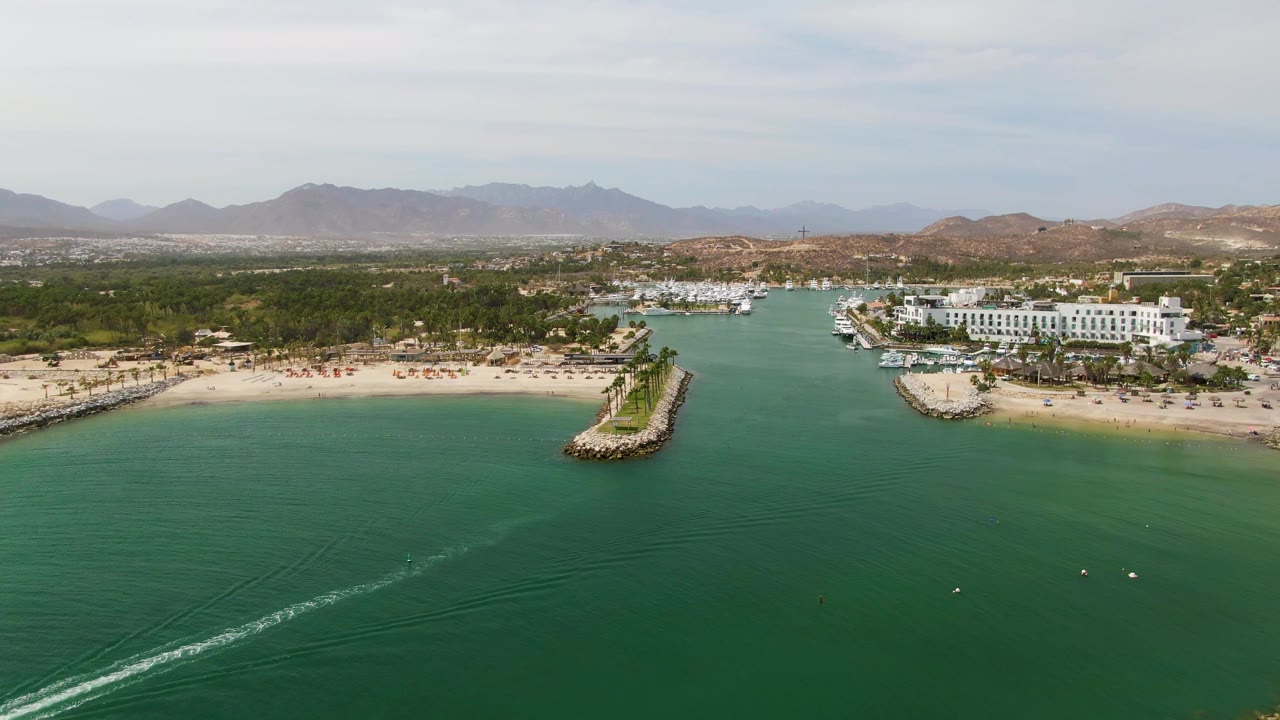 Aerial opening frame over Puerto Los Cabos Beach showing turquoise water, a sandy shore, a rocky breakwater, white resort buildings, marina boats, distant mountains, and no visible people.