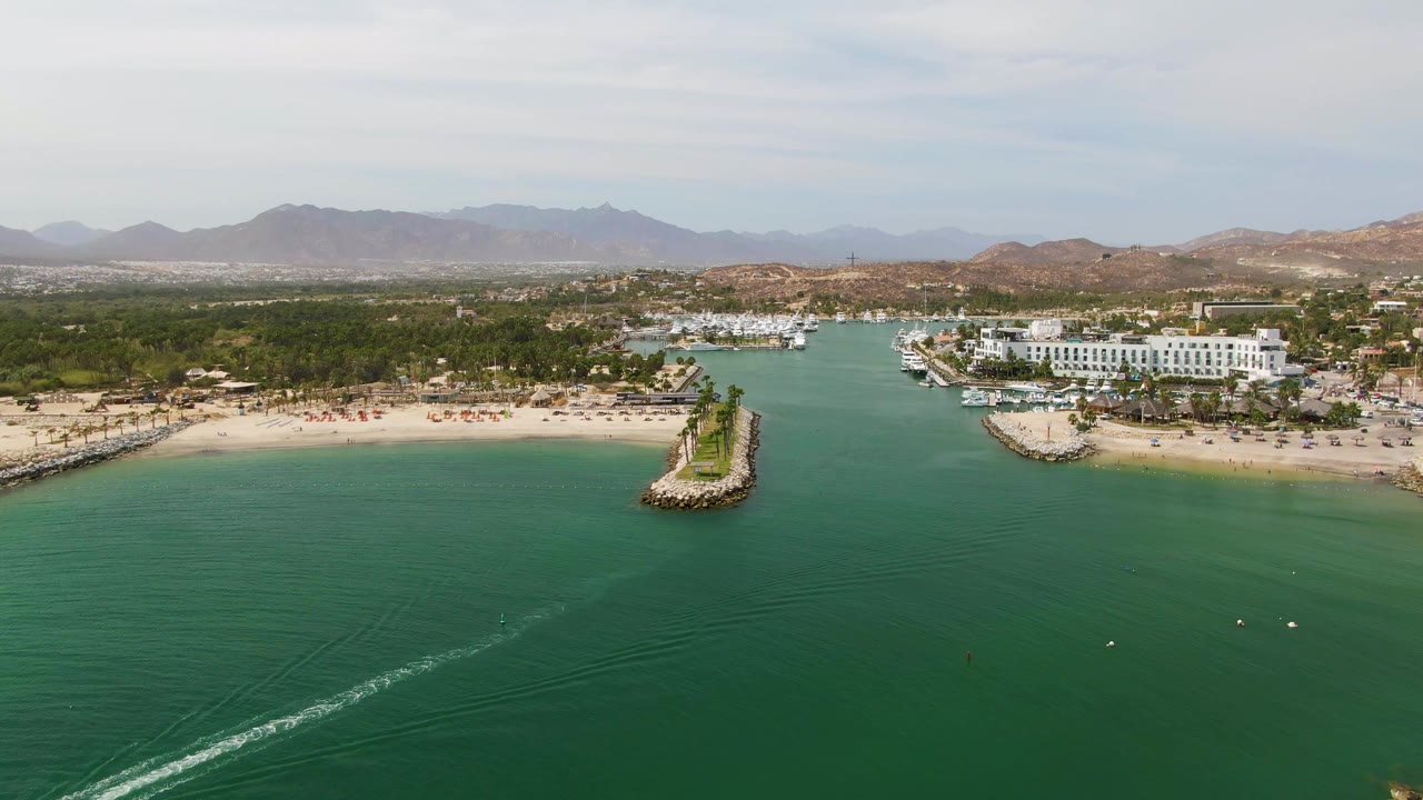 Wide aerial coastal view with a boat wake crossing green-blue water toward a sandy beach and marina.