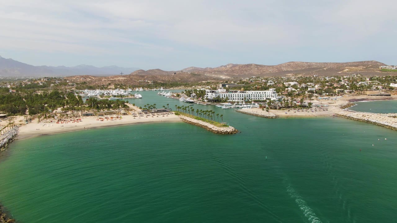 Opening aerial frame over Puerto Los Cabos Beach with turquoise water in front, a sandy beach and rocky breakwater through the middle, marina water and white buildings beyond, and no visible people.