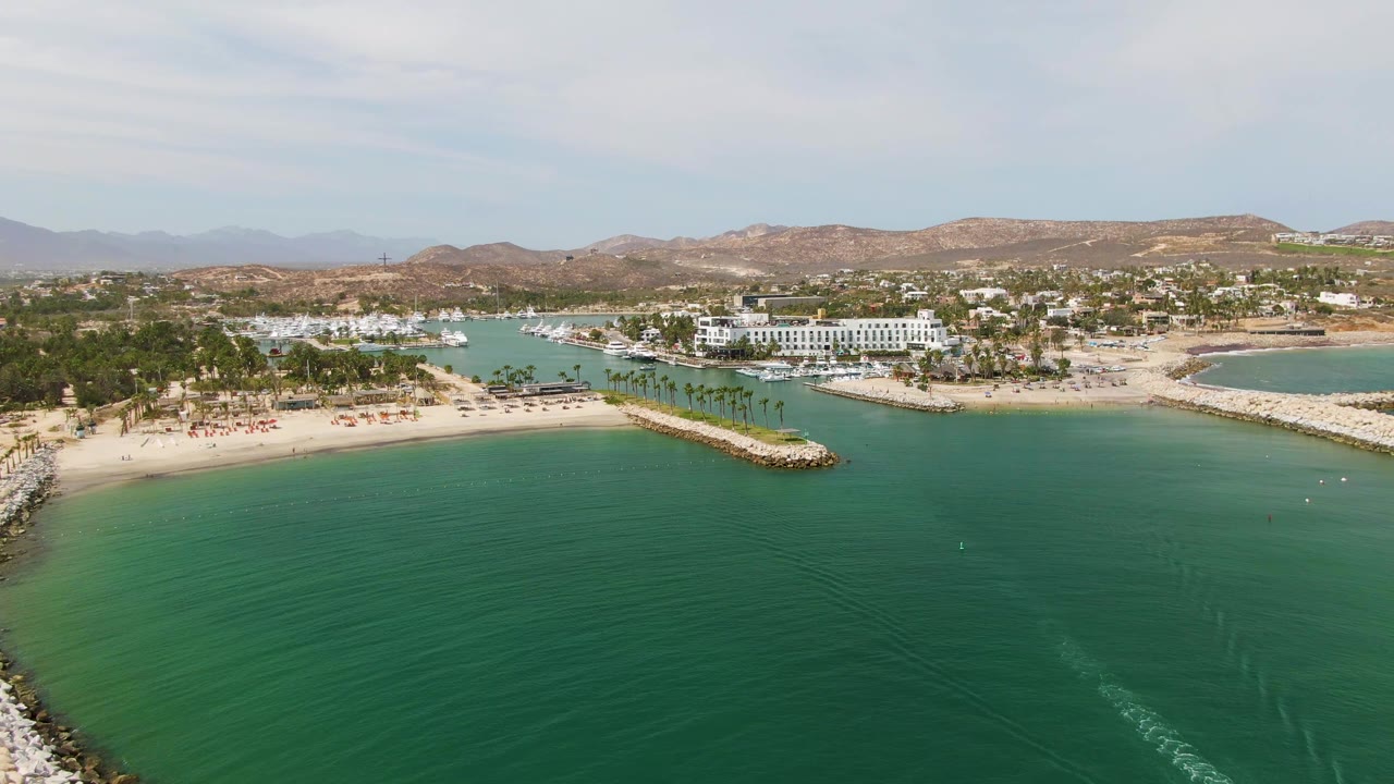 Drone view of curved breakwater beside calm turquoise water with beach, palms, marina, buildings, and hills.