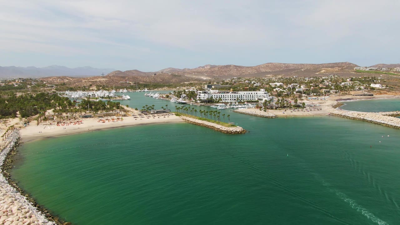 Puerto Los Cabos Beach appears in a wide elevated view with open turquoise water foreground, a pale rock breakwater along the sand, marina boats near white buildings, palm rows, brown hills, and no visible people.