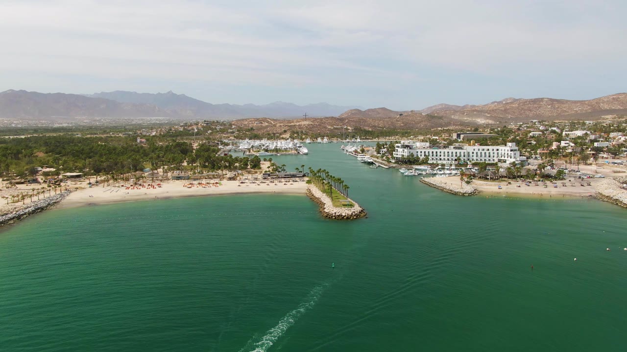 Wide aerial frame over Puerto Los Cabos Beach with turquoise water in the foreground, sandy shore and breakwater at center, marina buildings and mountains behind, and no visible people.