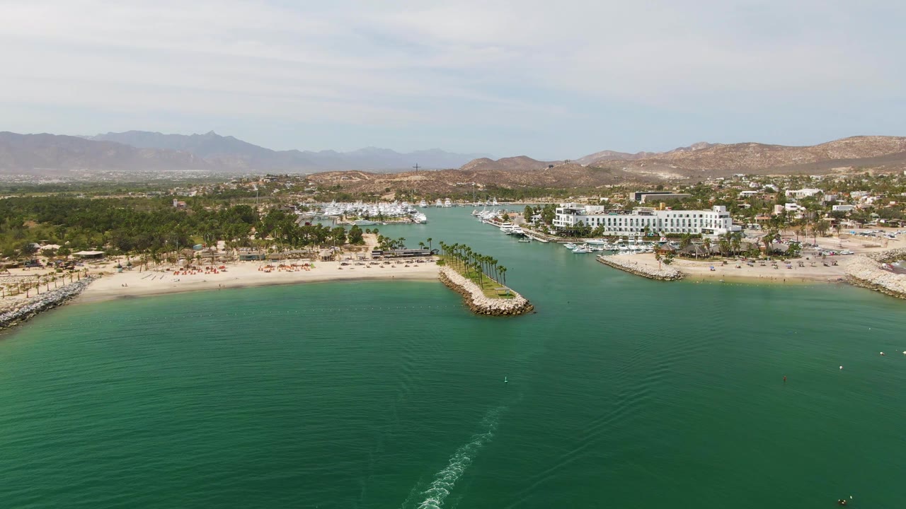 Puerto Los Cabos Beach aerial view with the boat wake beginning near the lower center, the sandy beach and palm-lined breakwater beyond it, and no visible people.