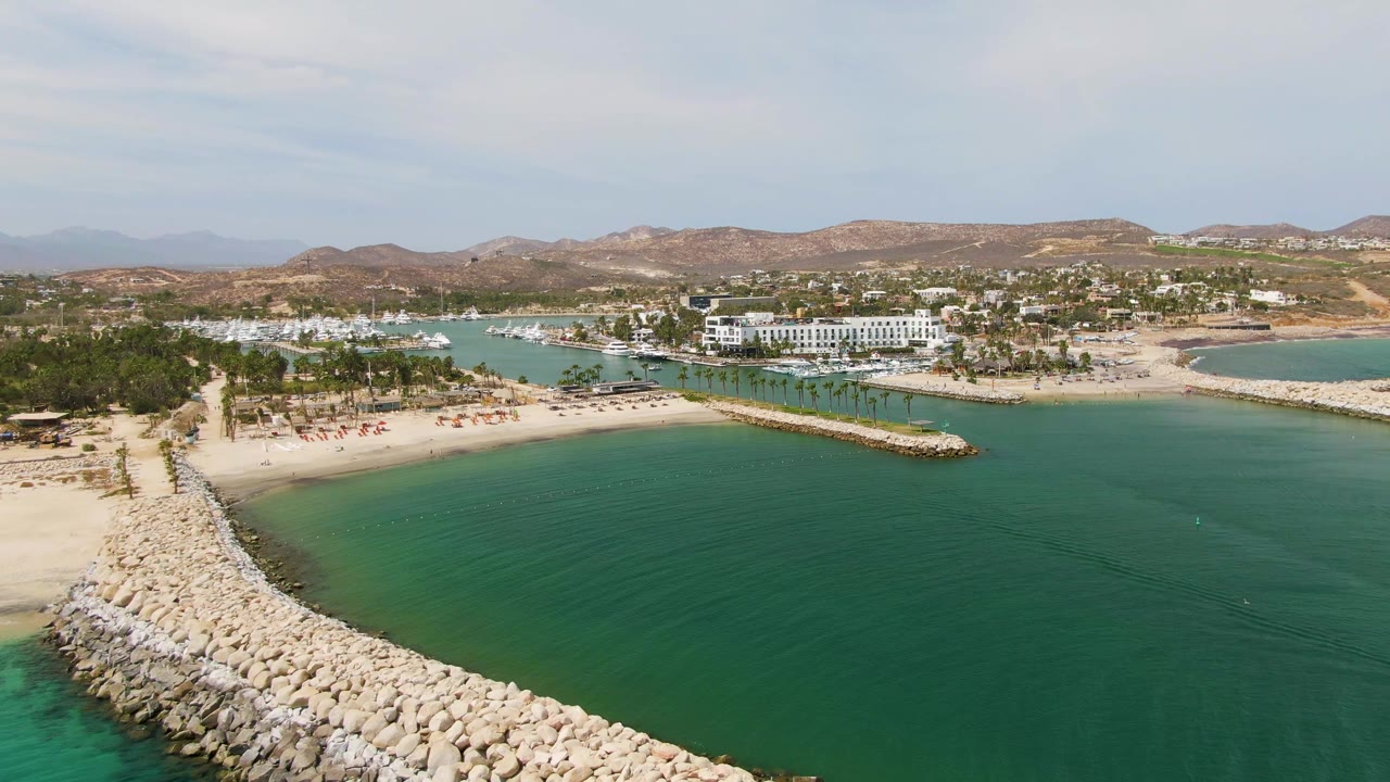 Aerial frame over Puerto Los Cabos Beach with a rocky breakwater entering from the lower left, turquoise water in front, sandy beach and palm trees beyond, and no visible people.