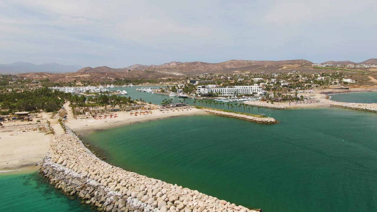 Aerial view at Puerto Los Cabos Beach with the rock breakwater extending across the lower frame, calm turquoise water beside it, beach umbrellas or small beach setups in the distance, and no visible people.