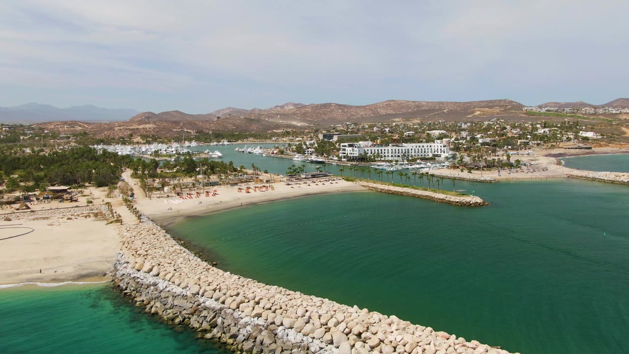 Aerial frame of Puerto Los Cabos Beach with the breakwater running from the bottom center toward the sandy shore, green-blue water on both sides, palm-lined beach area, and no visible people.