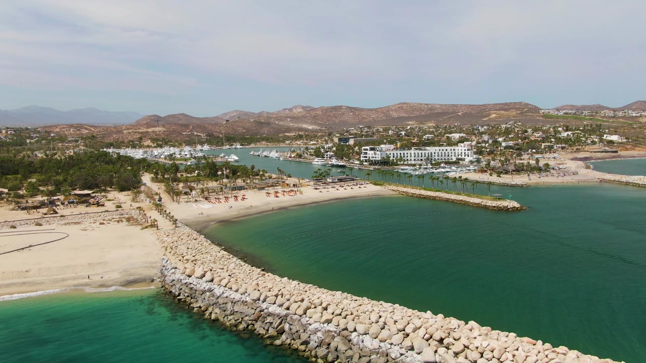 Wide aerial view of a sandy beach, breakwater rocks, calm water, palms, buildings, and mountains.
