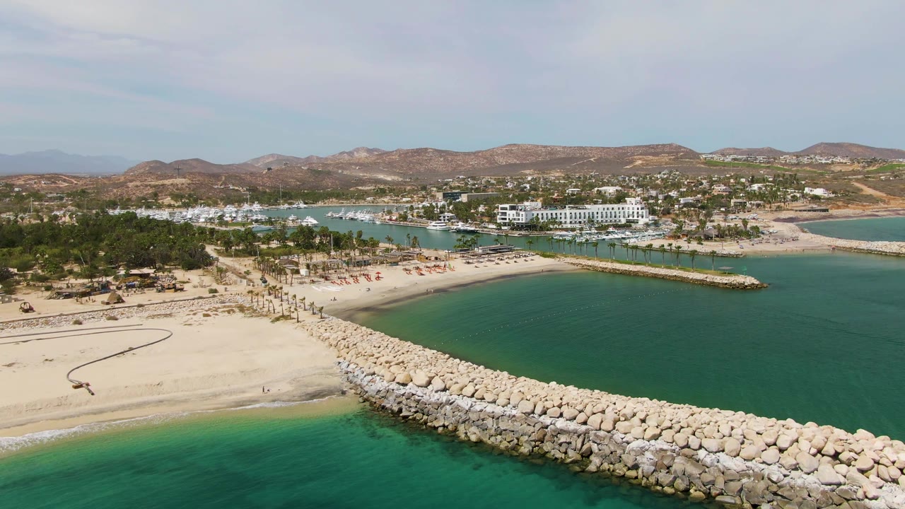 Aerial frame from Puerto Los Cabos Beach showing a pale rock breakwater crossing turquoise water beside broad sand, with palms, coastal buildings, and dry hills beyond.