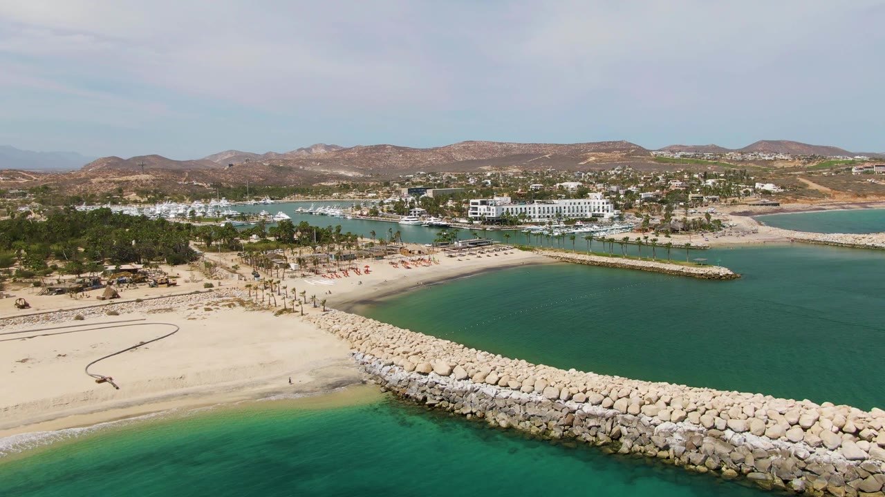 Wide aerial beach view with a diagonal rock breakwater, green-blue water, palms, buildings, and hills.