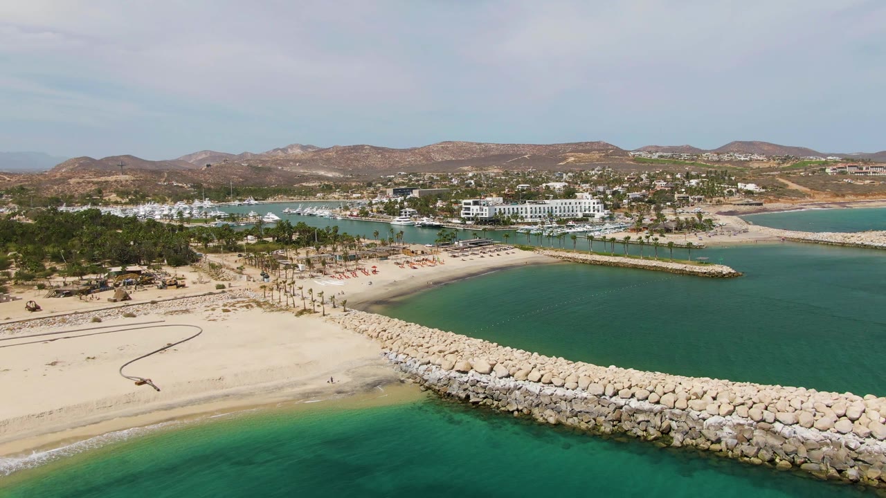 Aerial view at Puerto Los Cabos Beach with the rock breakwater stretching across the lower frame, a calm turquoise bay to the right, palm-lined sand to the left, and hillside development in back.