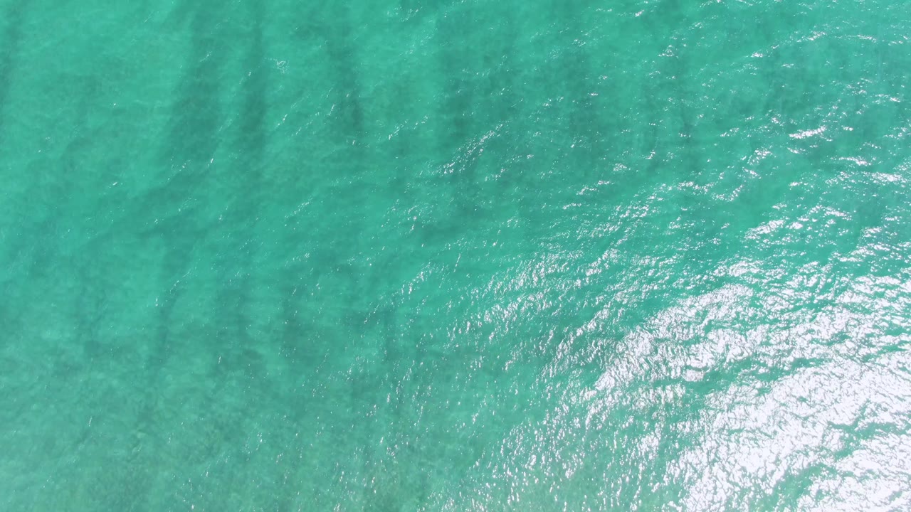 A brighter patch of turquoise water at Puerto Los Cabos Beach carries small sunlight glints over darker underwater streaks, with no visible people.