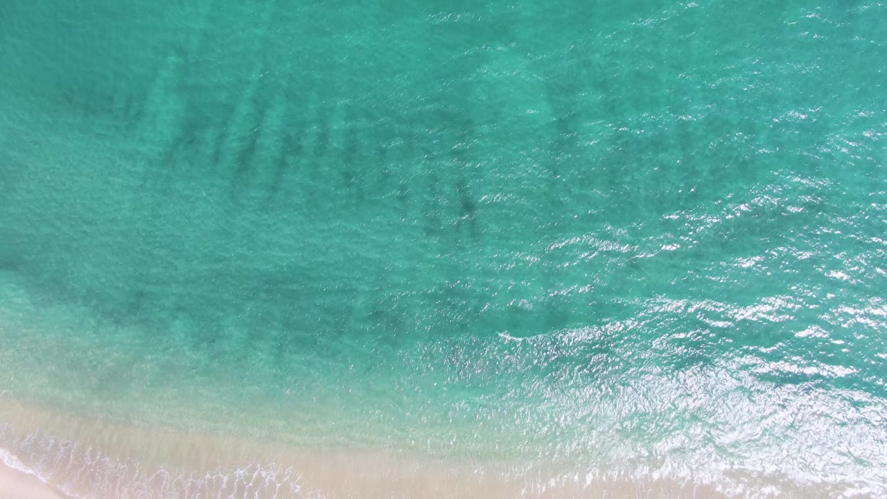 Clear turquoise water ripples over the seabed at Puerto Los Cabos Beach while soft white surf reaches the lower sand edge, with no visible people.