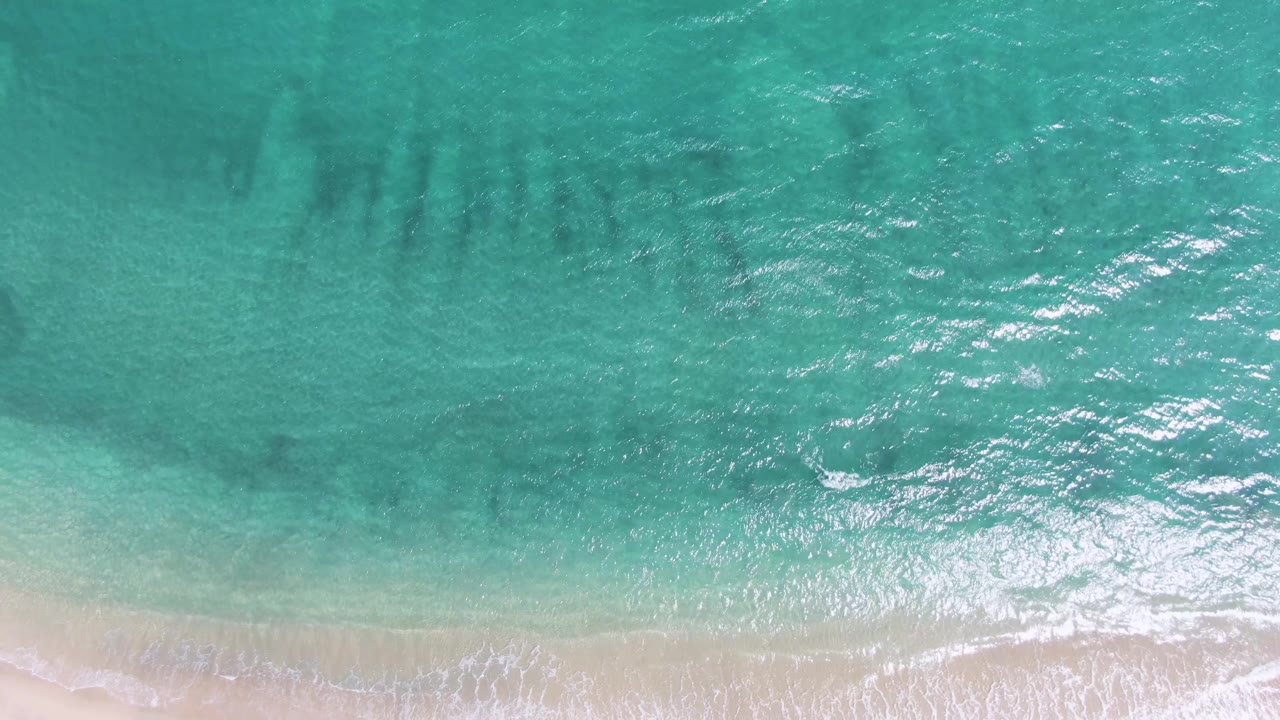 Sun reflections brighten the rippled turquoise water at Puerto Los Cabos Beach above a pale sand strip and thin foam band, with no visible people.