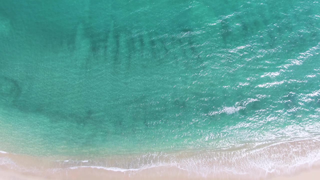 A wider lower band of pale sand appears beneath turquoise surf at Puerto Los Cabos Beach, with white foam tracing the shoreline and no visible people.