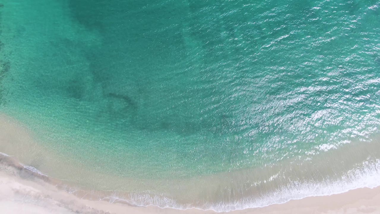Sunlit turquoise water covers most of the overhead Puerto Los Cabos Beach frame, with a thin white surf line meeting pale sand and no visible people.