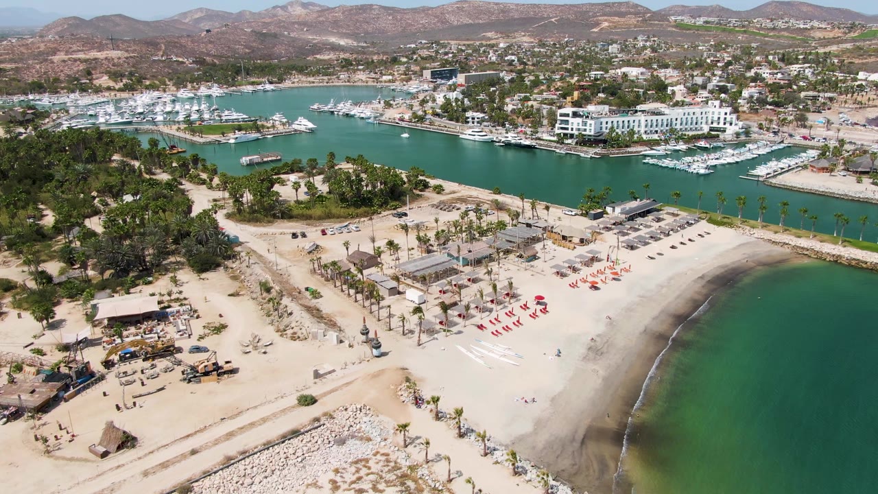 Aerial view of sandy beach, construction area, palm trees, marina, and turquoise water.