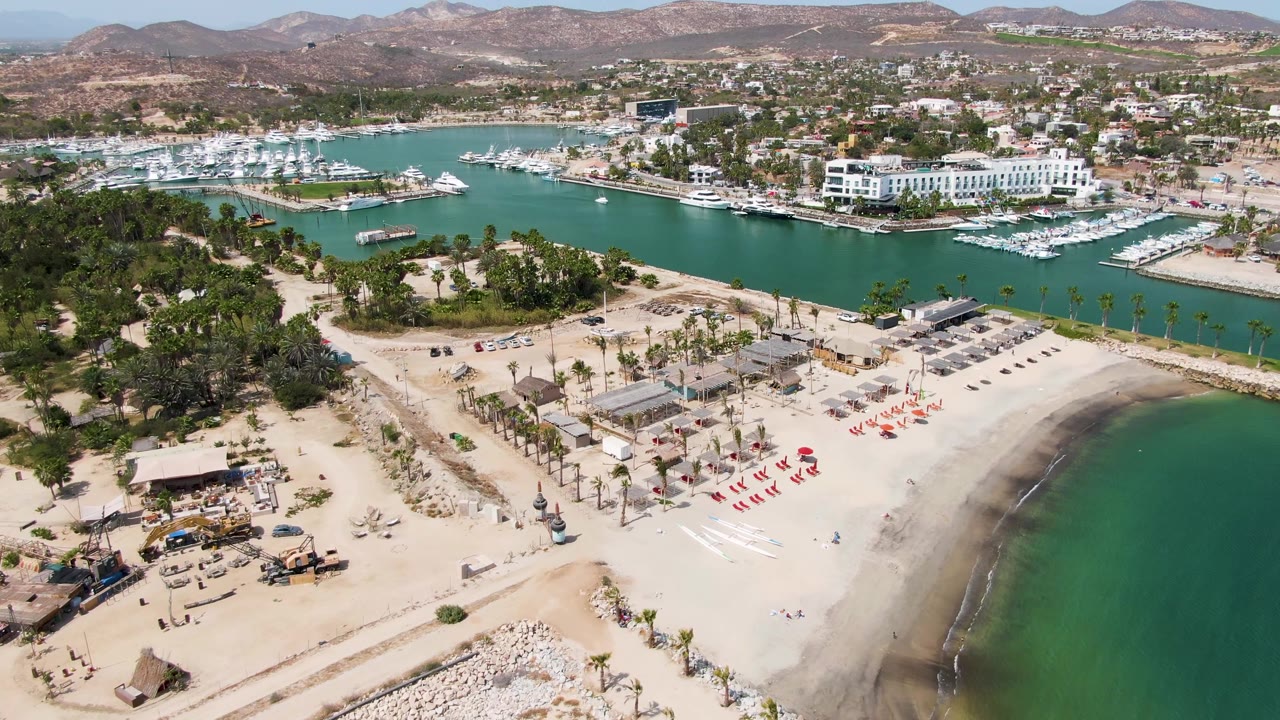 Puerto Los Cabos Beach from above with red chairs arranged on the sand, construction equipment near the lower left, green-blue marina water, docked boats, and no visible people.