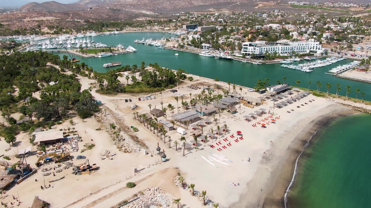 Puerto Los Cabos Beach aerial frame showing the curved shoreline at right, red lounge chairs on open sand, palms through the center, marina docks and boats behind, and no visible people.