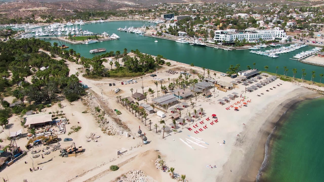 Puerto Los Cabos Beach drone view with construction materials and machinery at lower left, a palm-lined beach area, red lounge chairs, marina boats, white waterfront buildings, and no visible people.