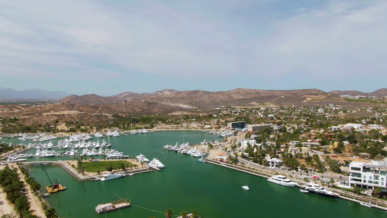 Aerial frame over Puerto Los Cabos Beach showing green marina water, docked white yachts, waterfront buildings, hillside homes, dry hills, pale sky, and no visible people.