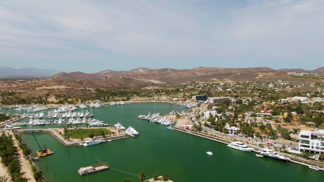 Wide aerial marina view with white yachts, green harbor water, waterfront roads, and hills.