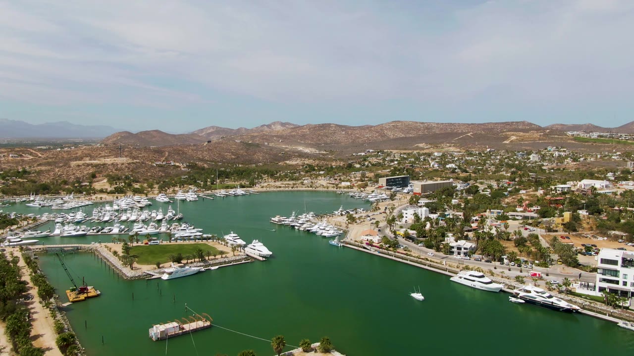Puerto Los Cabos Beach marina fills the frame with open green water in front, docked yachts to both sides, white waterfront structures, residential hills beyond, and no visible people.