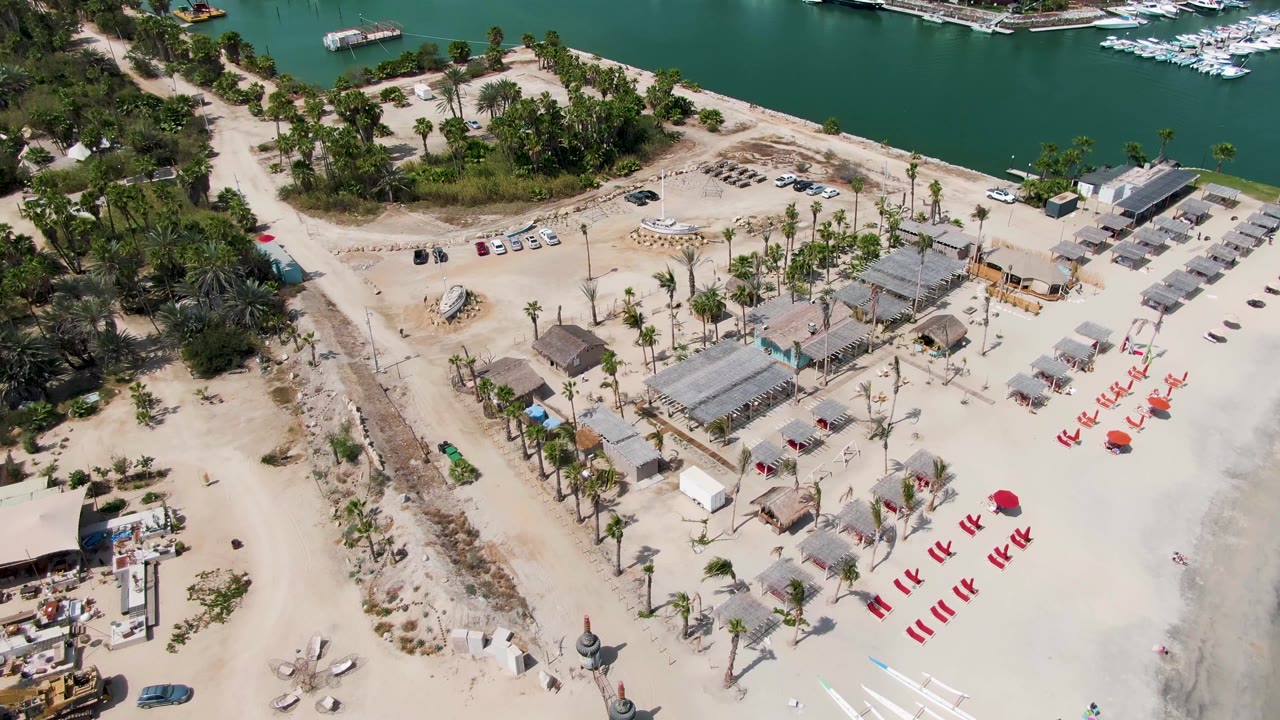 Drone frame over Puerto Los Cabos Beach showing clustered huts, a dirt access road, planted palms, red chairs on sand, marina water and boats in the background, and no visible people.