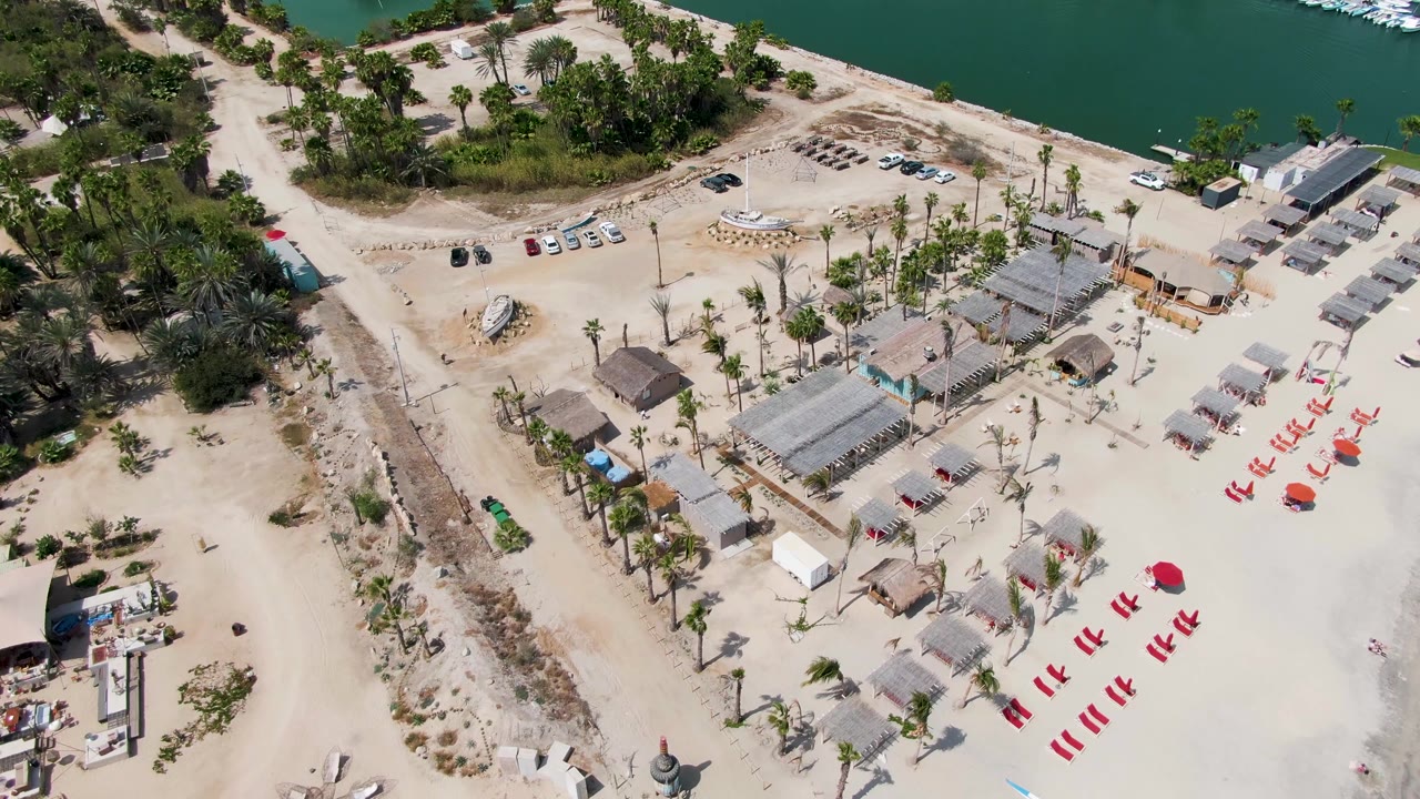 Closer overhead frame at Puerto Los Cabos Beach with huts and shade structures crossing the center, red beach chairs at right, dirt roadway at left, marina water near the top, and no visible people.