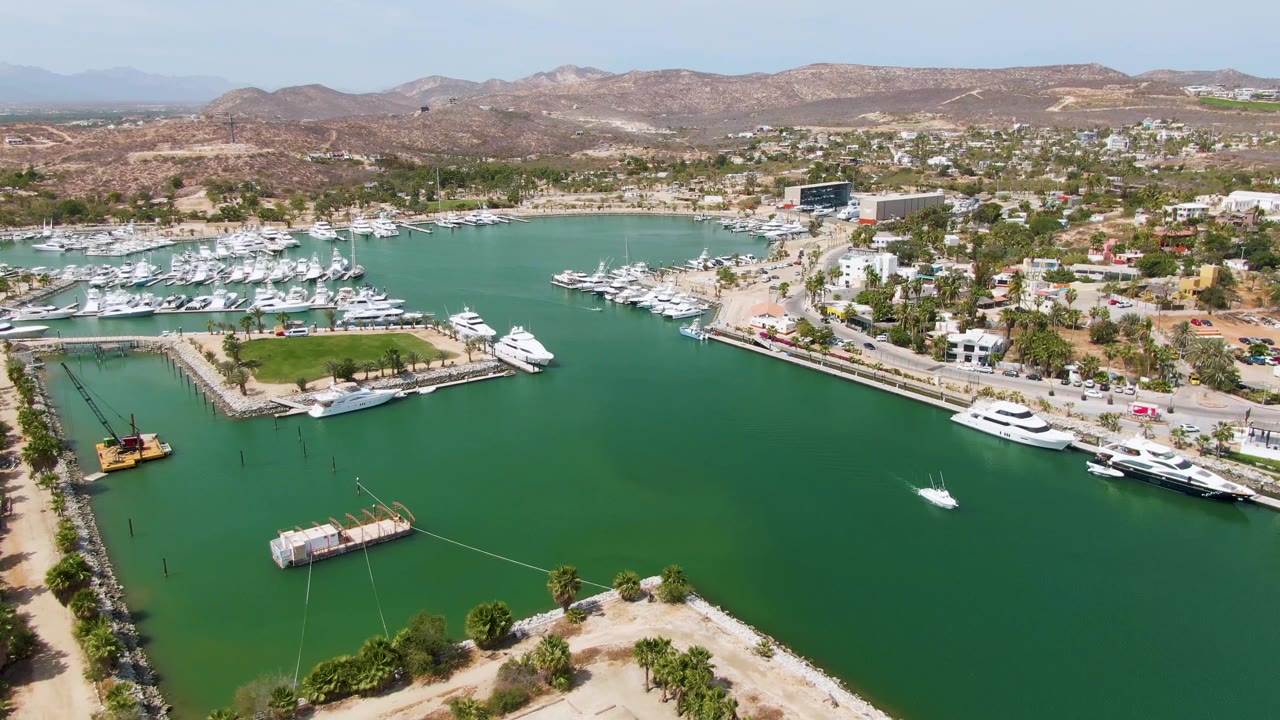Aerial view over Puerto Los Cabos Beach marina with green harbor water, docked yachts, a crane barge, shoreline roads, dry hills, and no visible people.