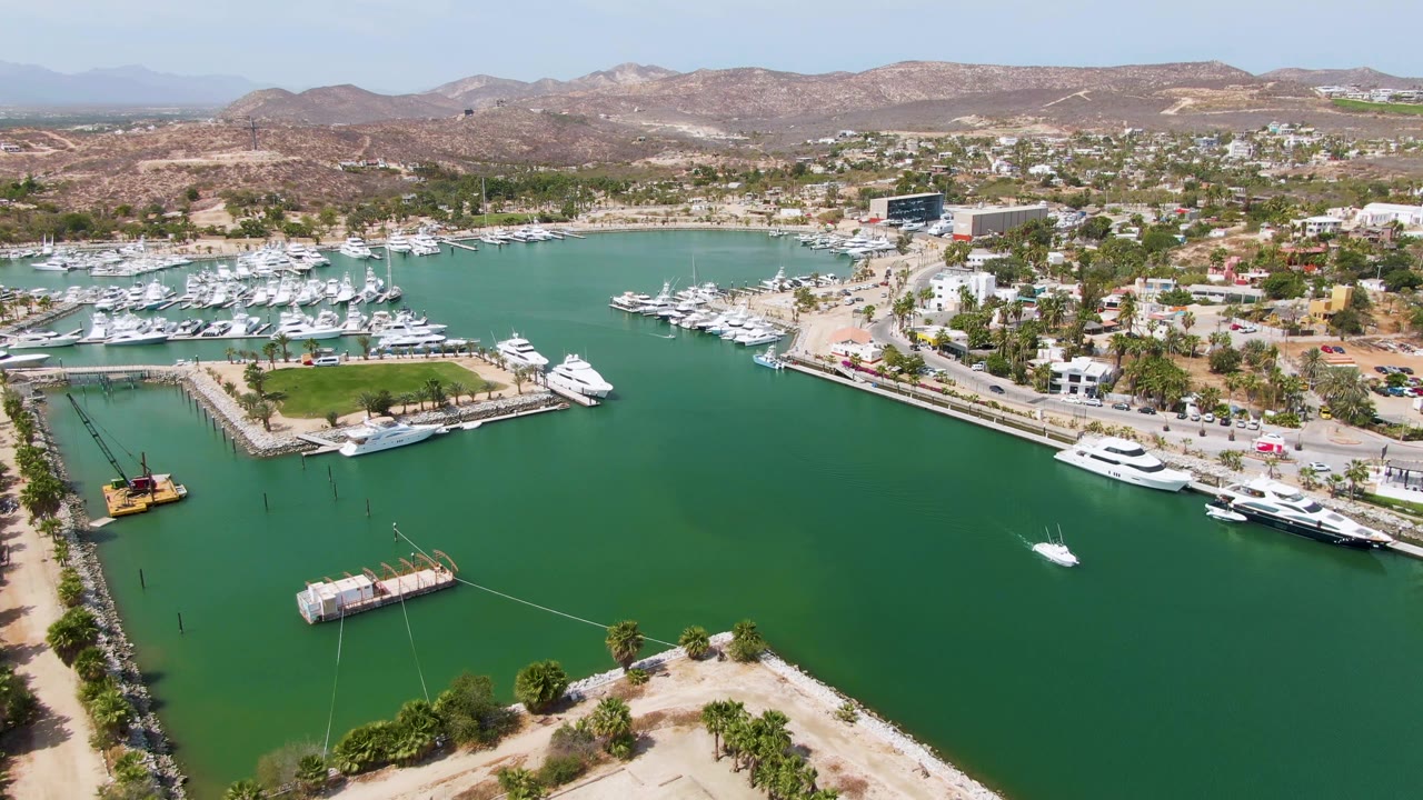 Aerial frame over Puerto Los Cabos Beach marina with dense rows of white yachts, a central road into the waterfront, green water, hillside terrain, and no visible people.