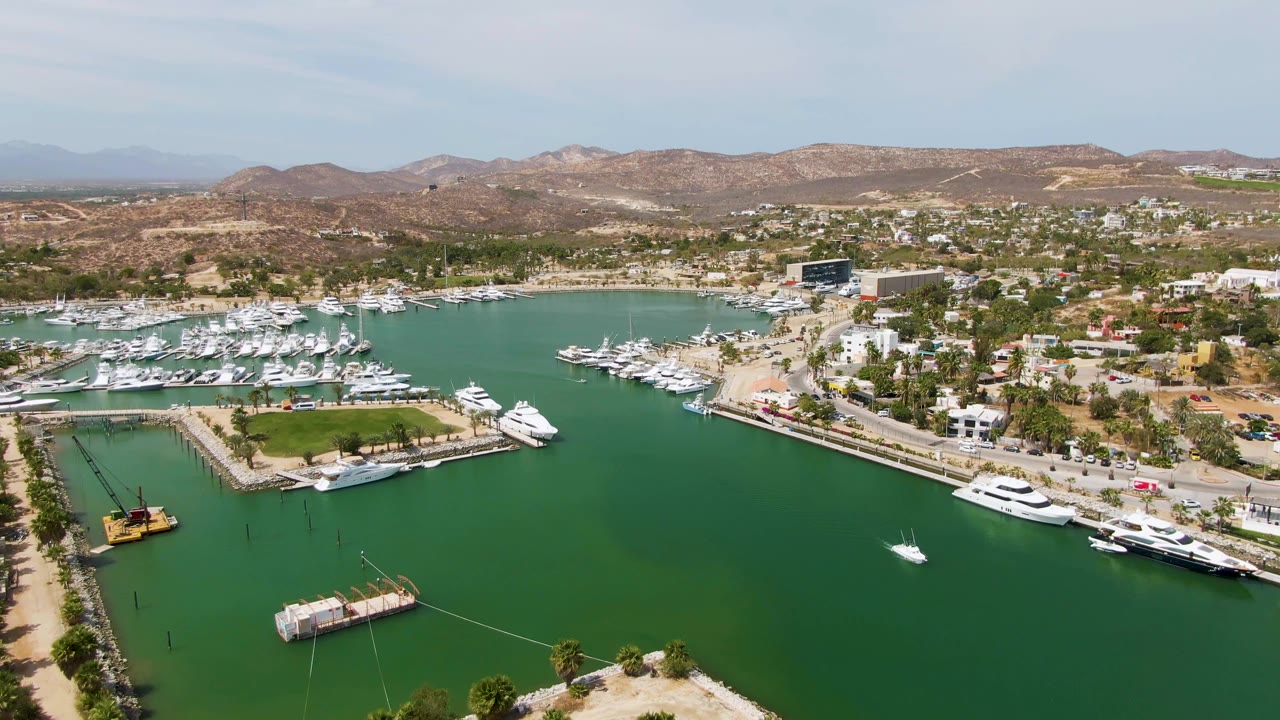 Second frame over Puerto Los Cabos Beach marina, with more shoreline road visible beside green water, rows of docked boats, hillside homes or buildings, and no visible people.