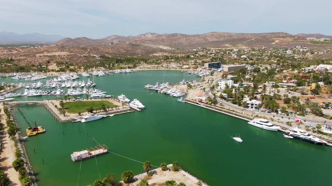Mid-clip aerial frame of Puerto Los Cabos Beach marina, with open green water in the foreground, docked yachts along the edges, clustered waterfront buildings, dry hills, and no visible people.