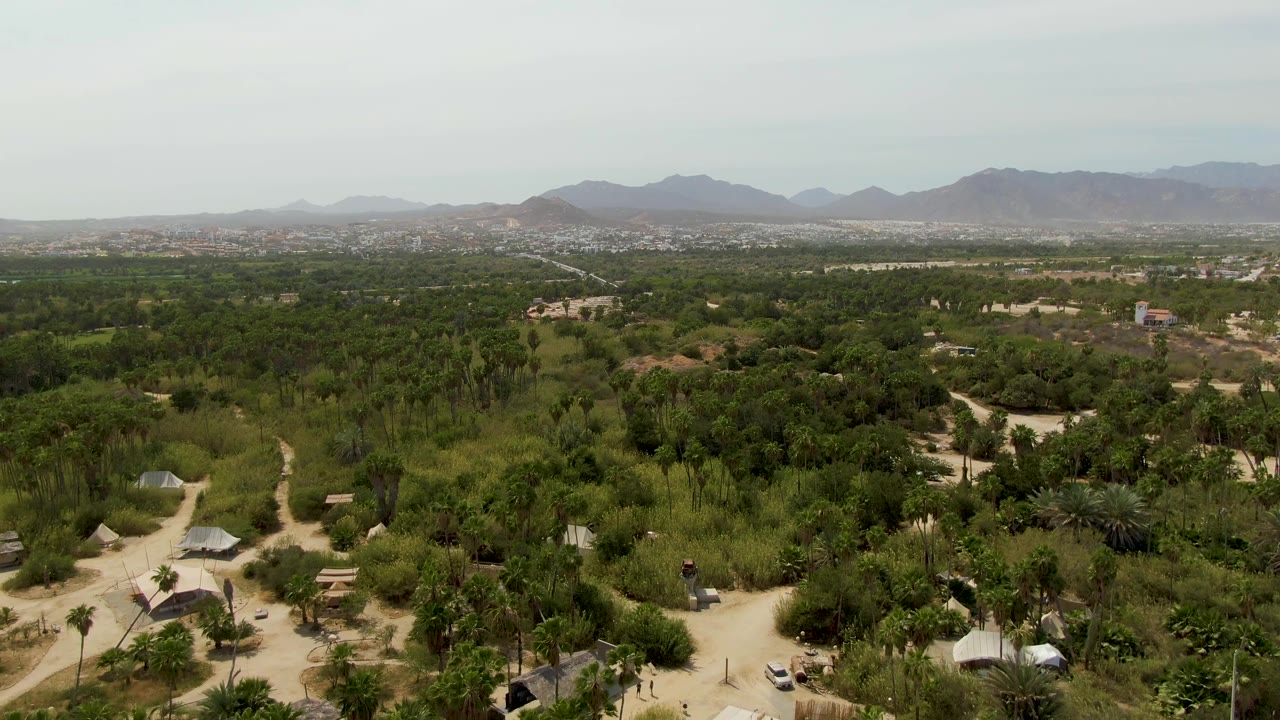 Wide aerial frame over Puerto Los Cabos Beach area vegetation, with pale paths weaving through huts and small buildings, mountains in the distance, and no visible people.