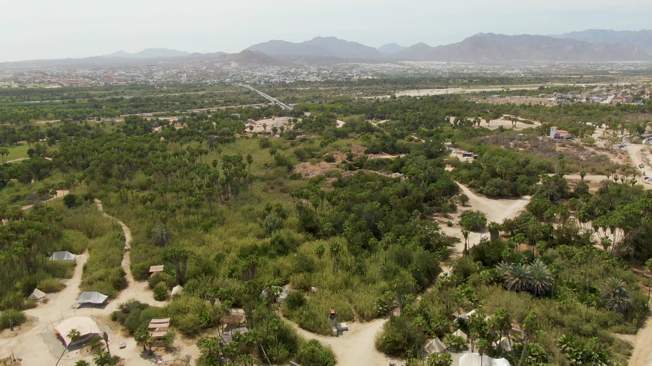 Aerial view over Puerto Los Cabos Beach, Baja California Sur, with pale dirt roads crossing dense greenery, scattered small structures, hazy mountains on the horizon, and no visible people.