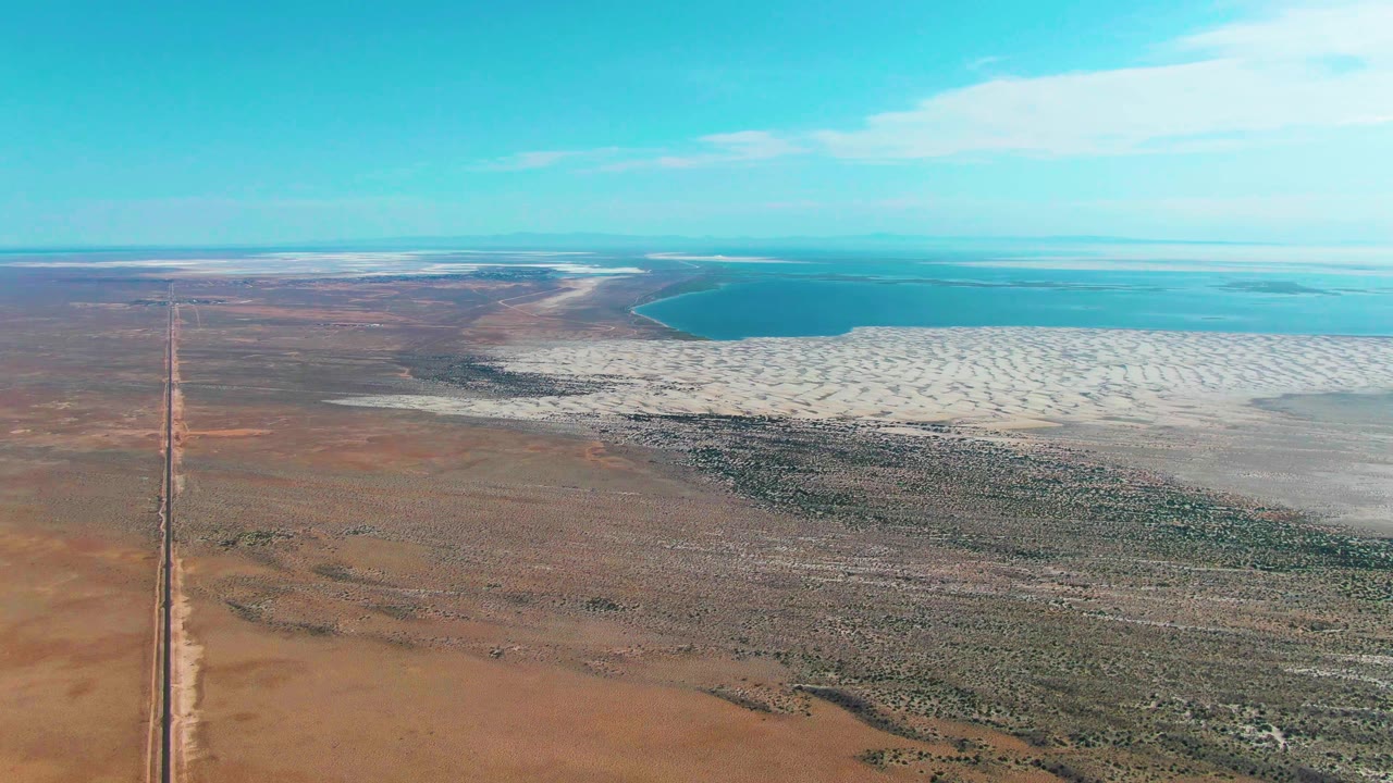 Aerial view of a dirt road beside desert scrub, white salt flat, and blue water.