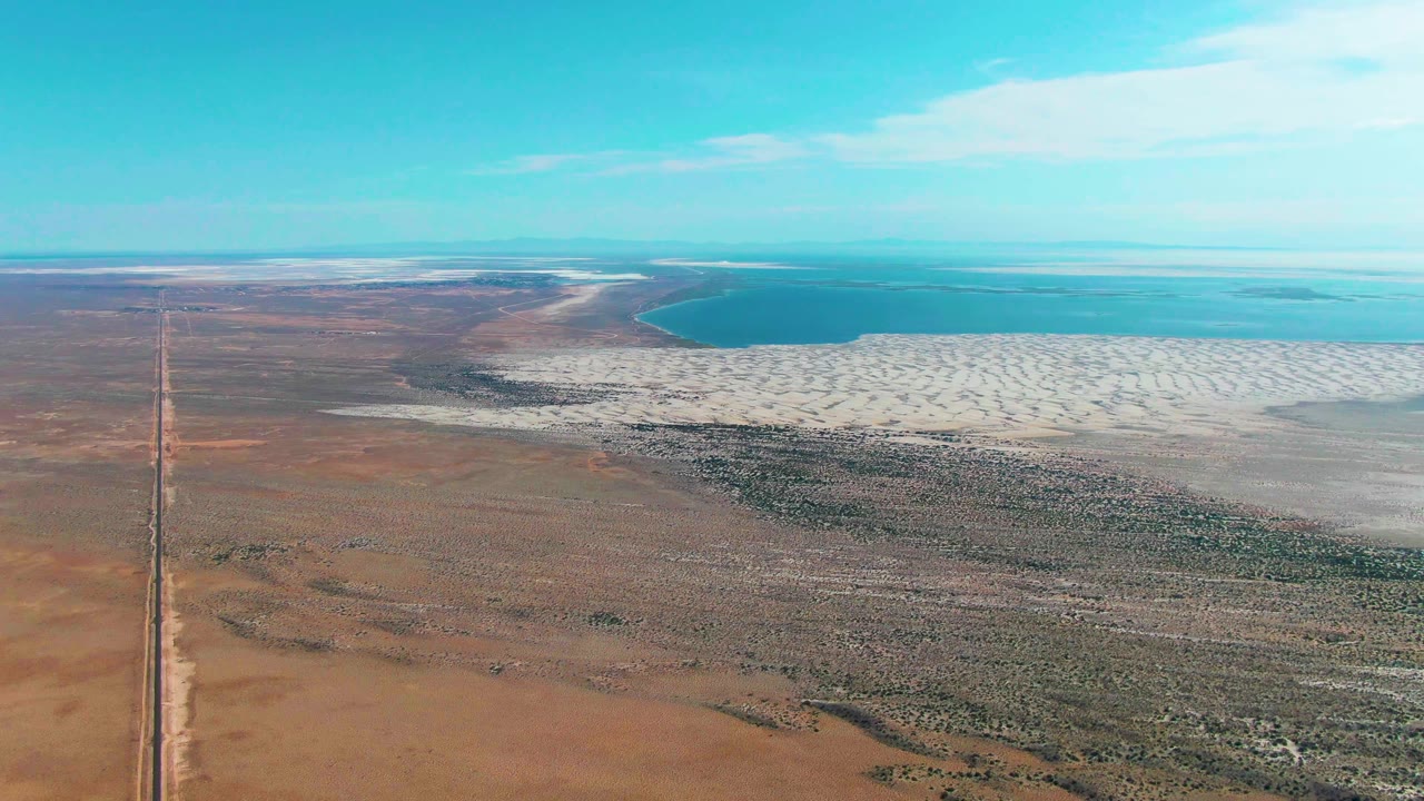 One second in, the Guerrero Negro aerial view keeps the dirt road near the left edge while the white salt flat spreads across the right toward blue water, with no visible people.