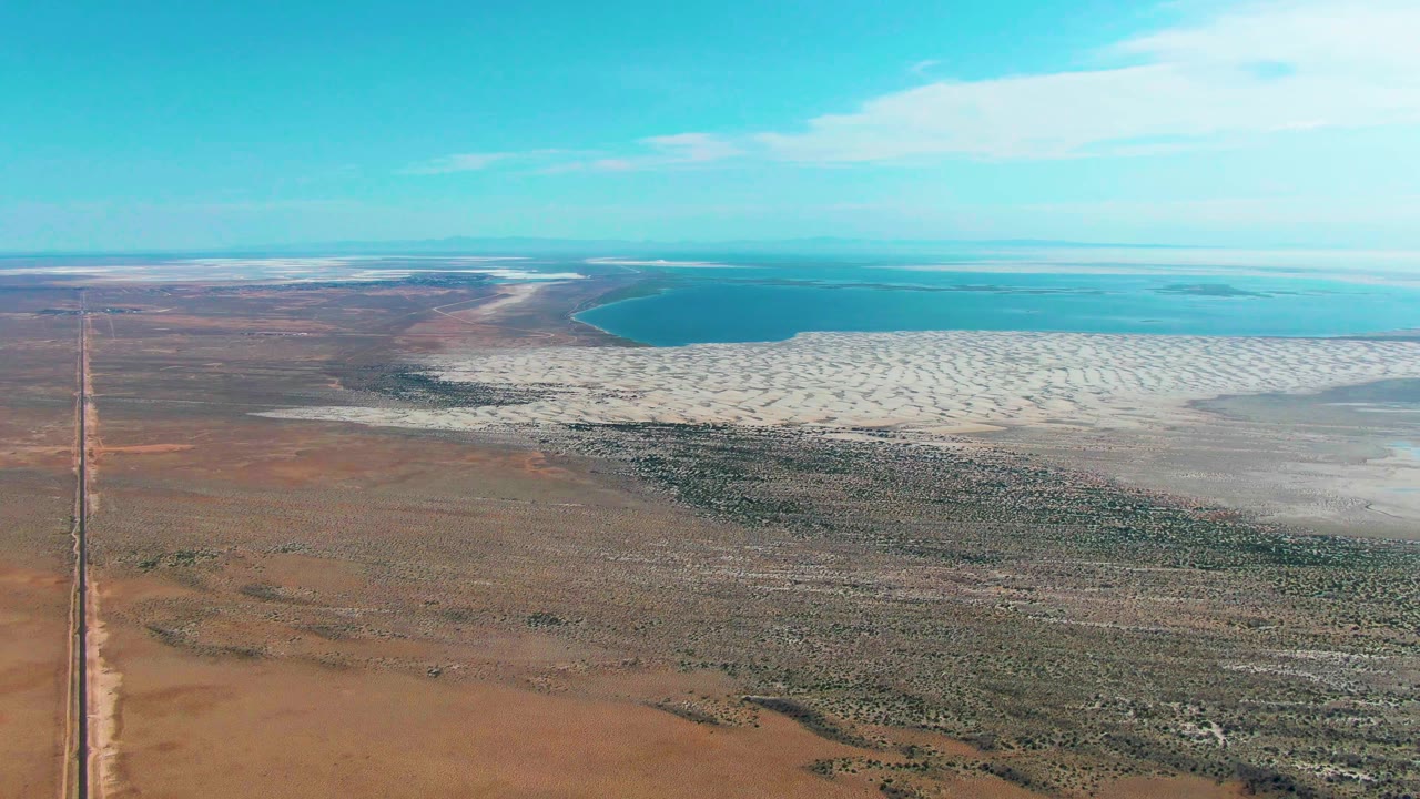 Aerial frame over Guerrero Negro, Baja California Sur, showing brown arid ground in front, a white salt flat beside blue water, a straight road at the far left, clear sky, and no visible people.