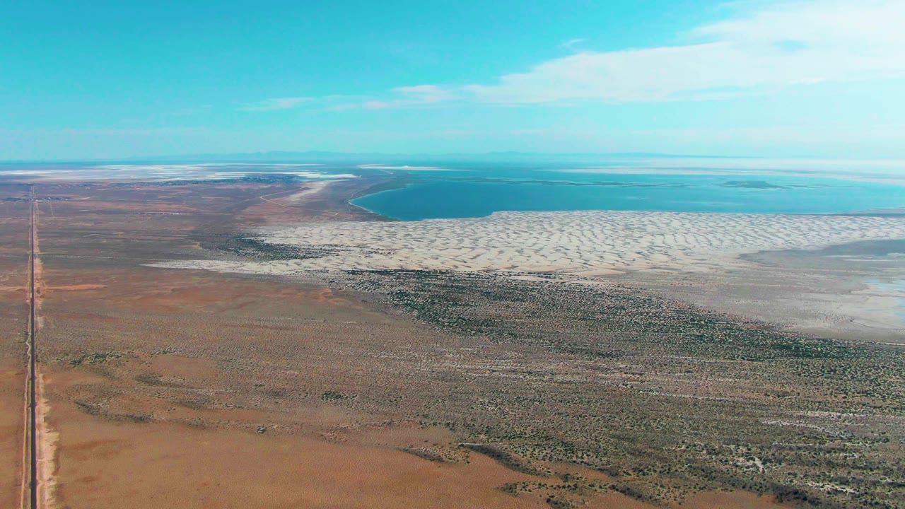 Aerial view in Guerrero Negro with the left-side road still visible, brown desert foreground, pale salt flat across the midground, blue water beyond, and no visible people.
