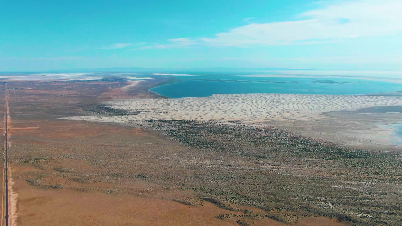 High aerial frame over Guerrero Negro showing brown arid land, a broad white salt flat, blue-green water, the left road near the edge, soft cloud cover, and no visible people.
