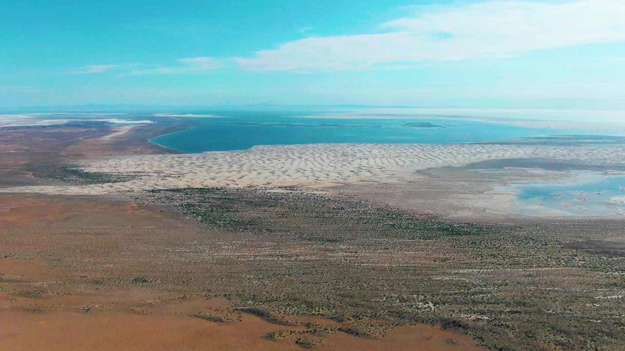 Wide drone view over Guerrero Negro showing a blue coastal inlet at left, pale sand and flats across the center, and sparse desert vegetation in the foreground, no visible people.