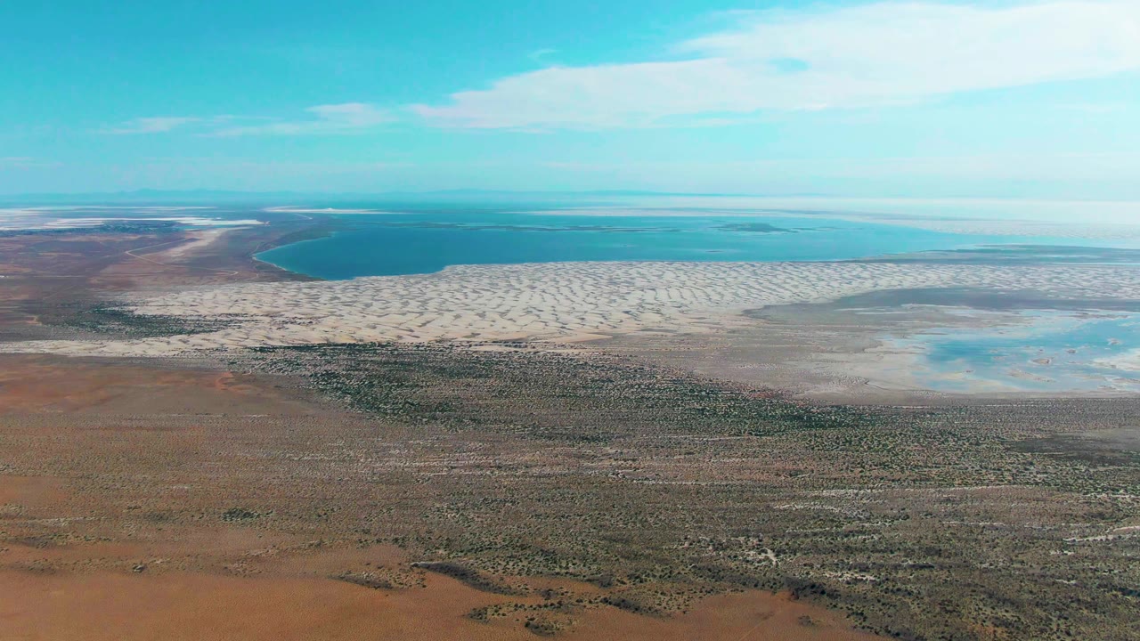 High aerial frame in Guerrero Negro with the ocean band running across the top, light sand flats below it, and mottled brown-green desert scrub filling the lower frame, no visible people.