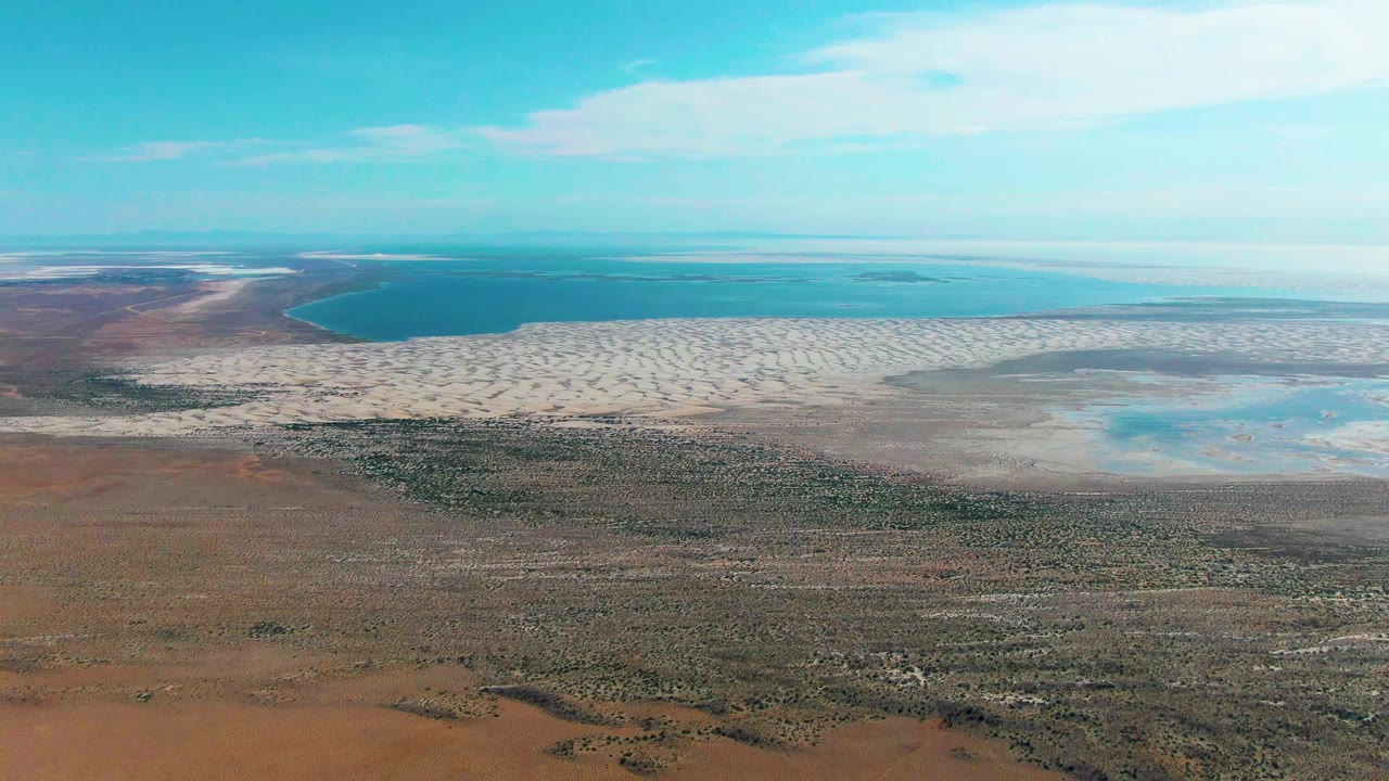 Aerial view over Guerrero Negro where turquoise water and a curved sandy edge sit above a broad field of dry brown land and low vegetation, no visible people.