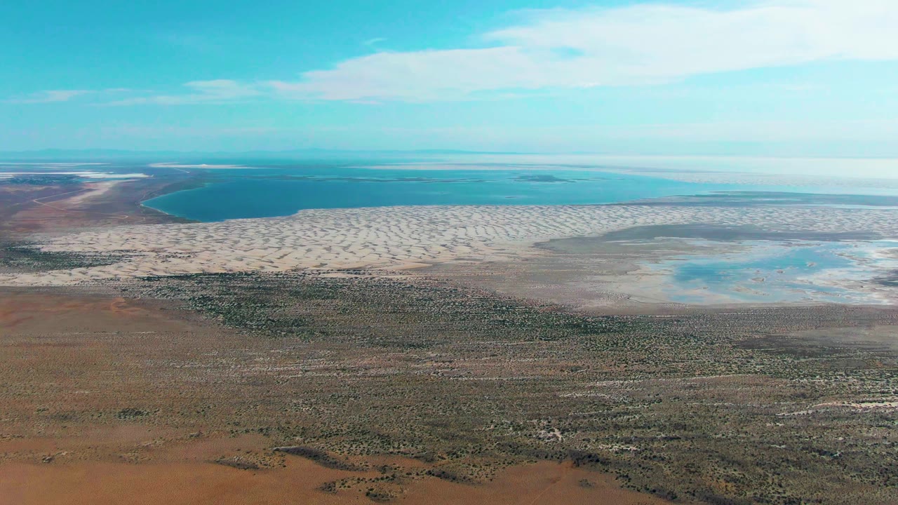 A high aerial frame over Guerrero Negro, Baja California Sur shows brown scrubland in the foreground, pale exposed shoreline flats, blue ocean beyond, thin clouds, and no visible people.