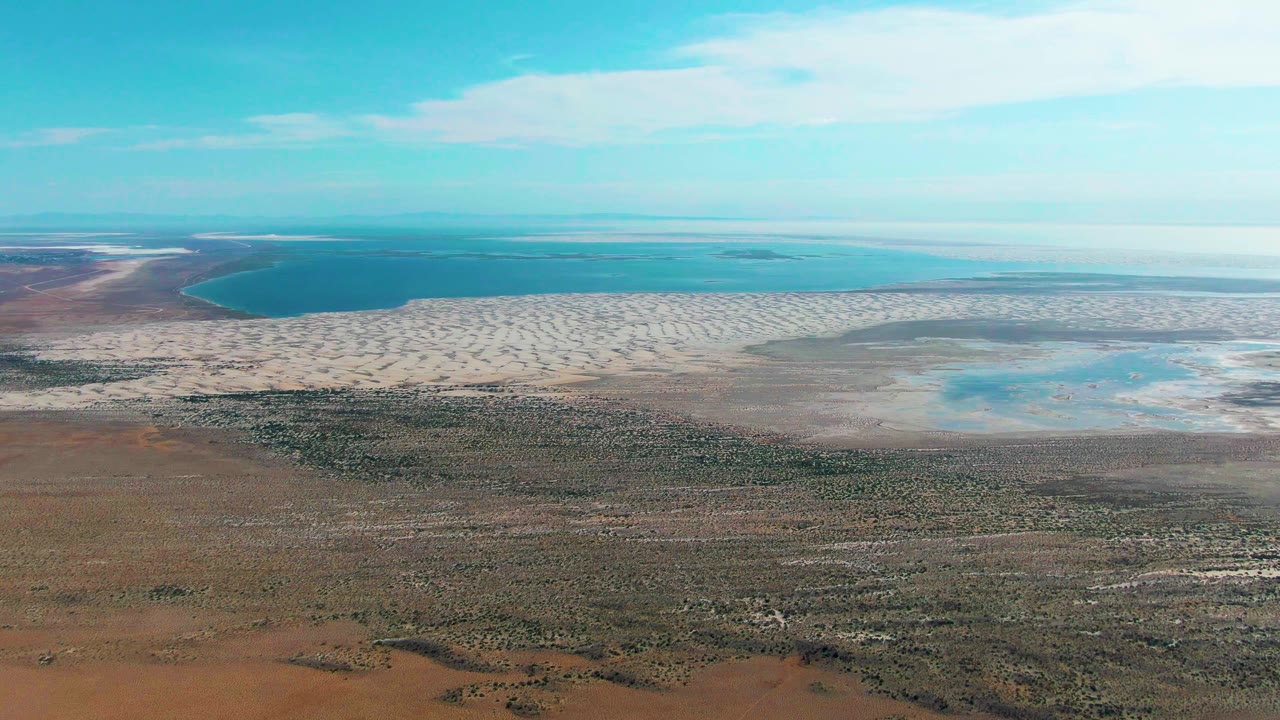 The 0:01 frame shows Guerrero Negro's dry vegetated foreground, a broad light-colored shoreline band, turquoise water spreading across the upper frame, and no visible people.