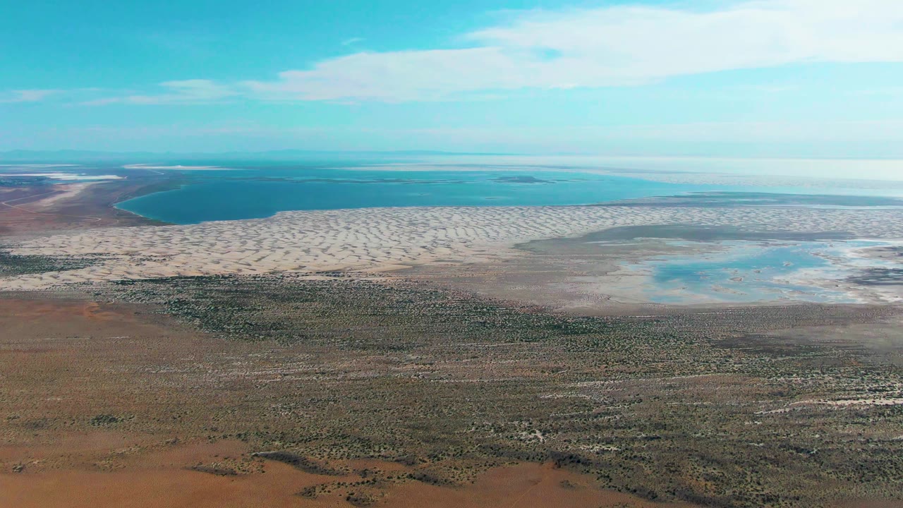 At 0:02, the aerial view over Guerrero Negro keeps the scrubby brown foreground low in frame, pale flats across the center, blue coastal water behind them, and no visible people.